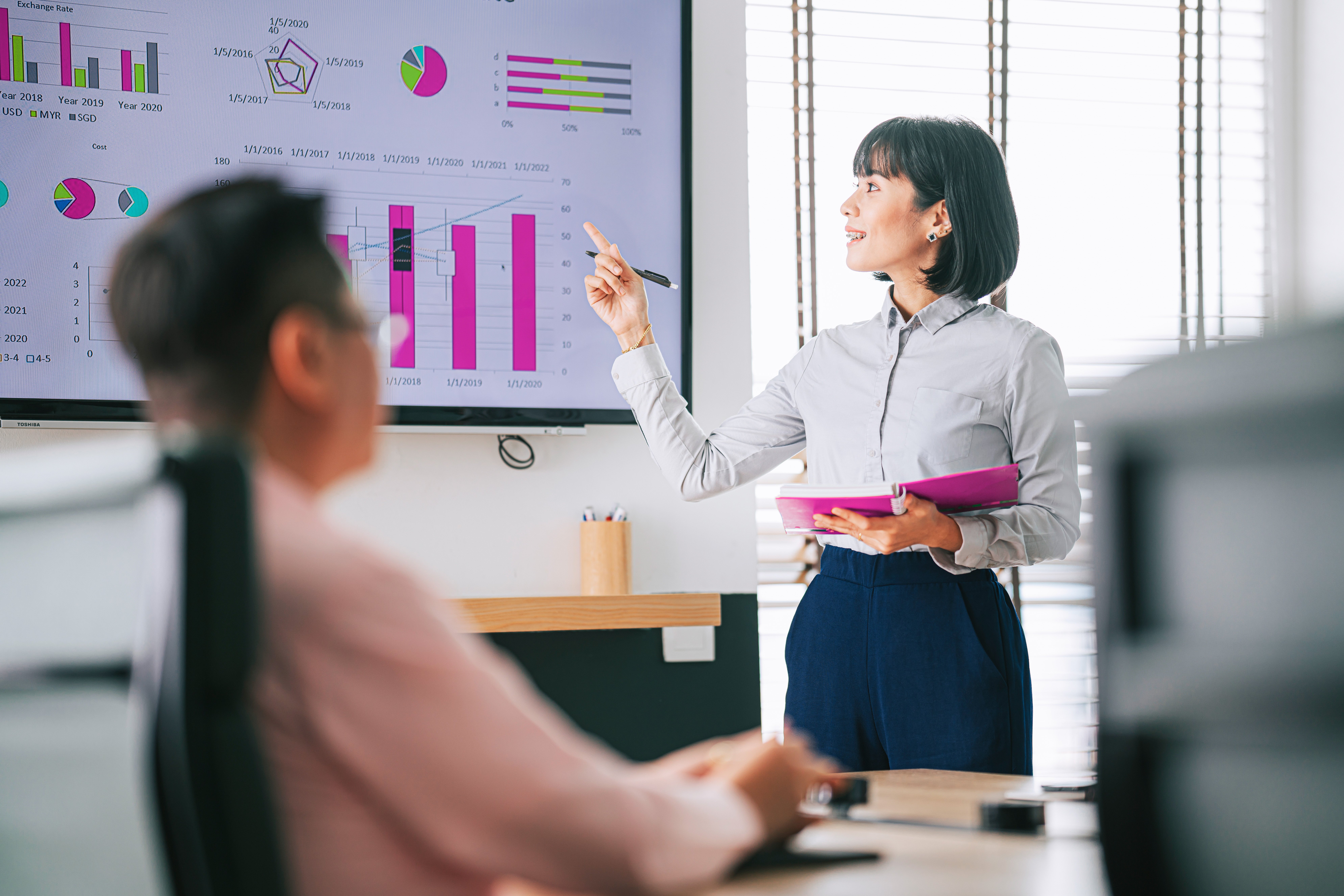 Asian Malay woman with dental braces confidently presenting to her colleague in conference room with television screen presentation