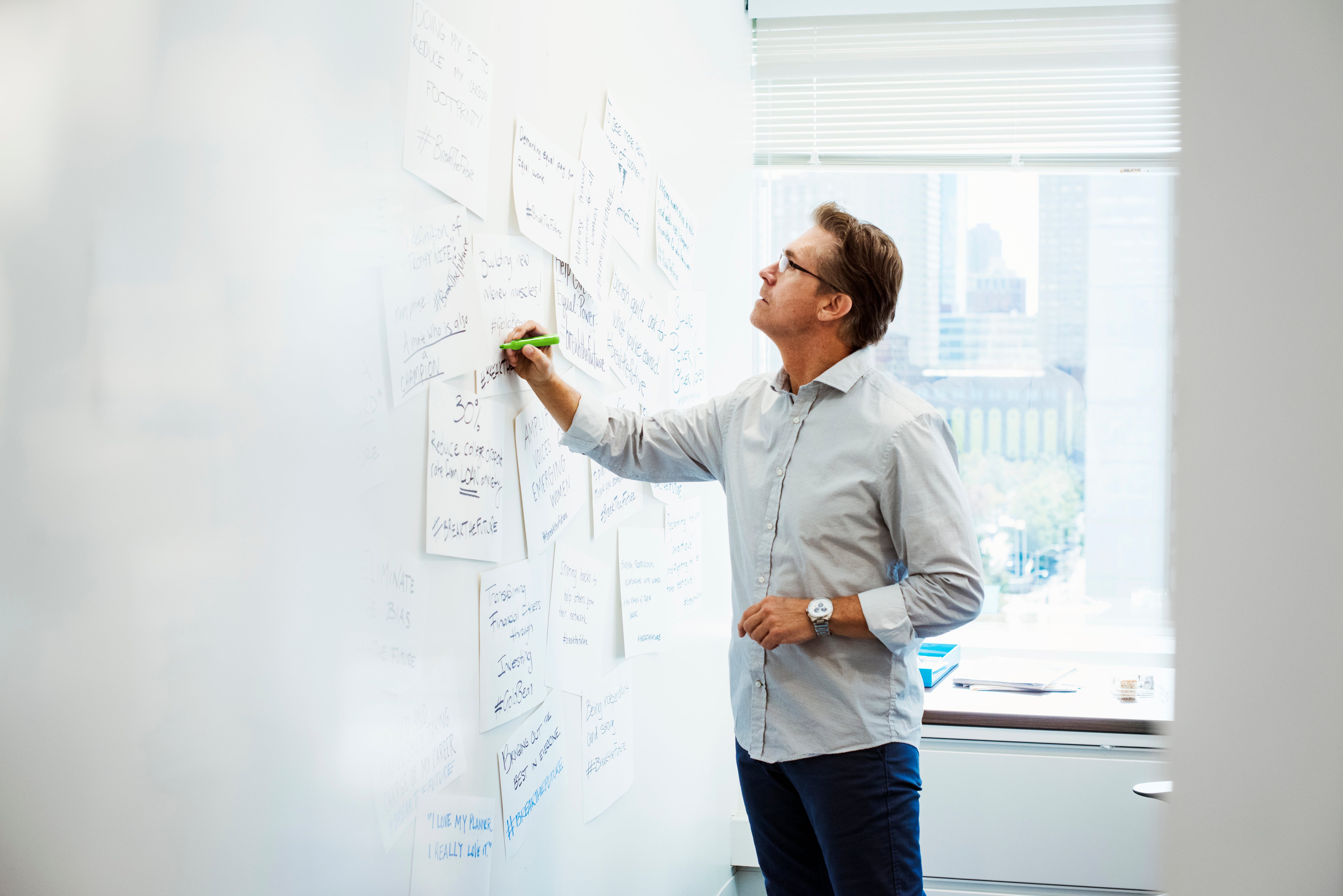 A man standing in an office writing on pieces of paper pinned on a whiteboard.
